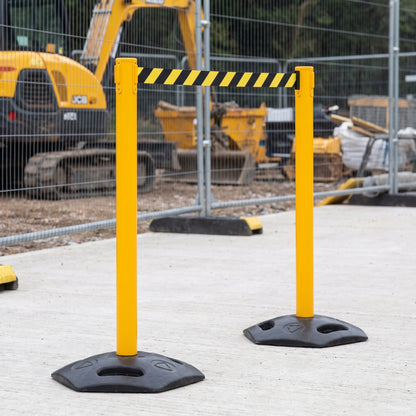 Yellow metal construction barrier posts with black bases and a black-and-yellow striped tape section, set on a concrete surface at a construction site.